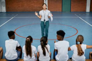Photo of Latina PE Teacher giving instructions to a group of diverse elementary Physical Education students.