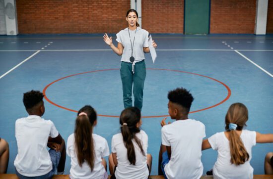 Photo of Latina PE Teacher giving instructions to a group of diverse elementary Physical Education students.