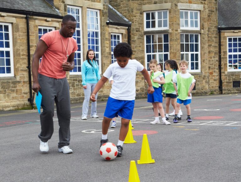 Black male PE teacher coaching a group of diverse elementary PE students through a soccer/foot dribbling exercise weaving between yellow cones.